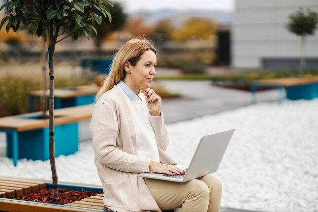 A focused businesswoman is thinking about report while sitting in a park downtown with a laptop