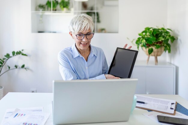 Focused businesswoman presenting charts and graphs on video\
call online. business, technology and people concept, mature\
businesswoman with tablet having video conference by laptop\
computer at office