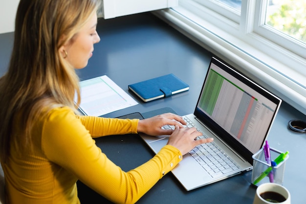 focused caucasian woman sitting at desk working from home and using laptop in sunny room