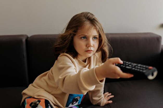Focused charming little girl sitting on sofa with TV remote and searching for movie