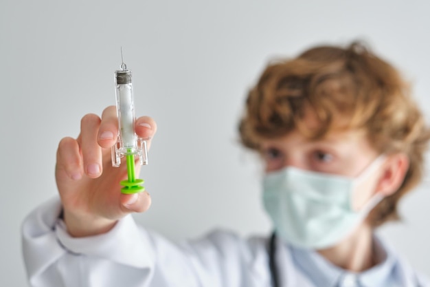 Focused child in disposable mask and medical uniform with plastic syringe on white background