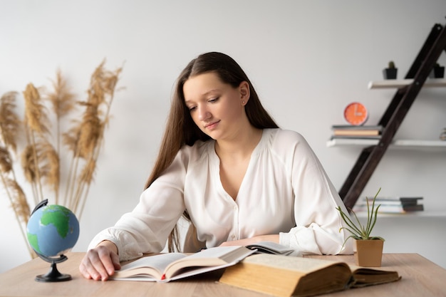 Focused college student sitting at his desk with open textbooks doing homework while studying at home Beautiful teenage Caucasian girl reads books in the library Education and knowledge concept