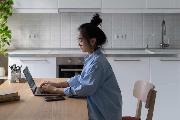 Focused concentrated young asian female freelance blogger working on laptop in kitchen