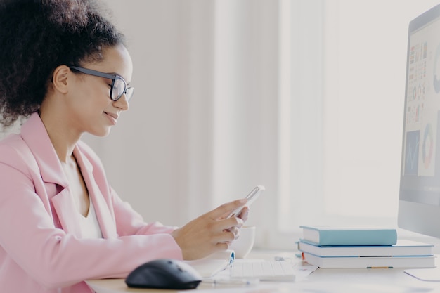 Focused curly haired female office worker holds mobile phone, searches information in internet