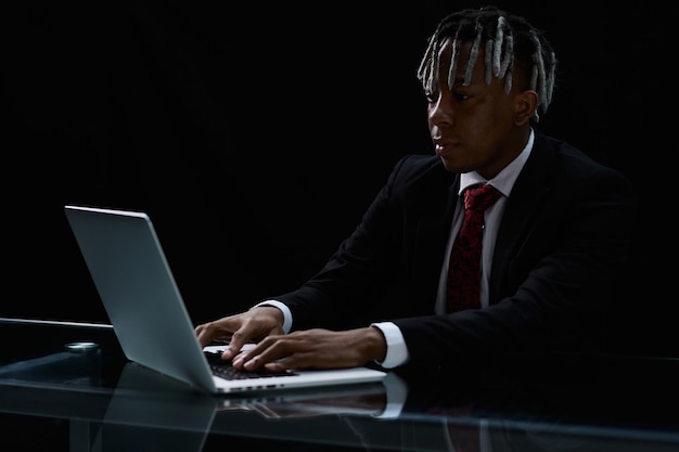 어두운 피부색의 사업가가 노트북을 사용하는 현대적인 사무실의 테이블에 앉아 있습니다.