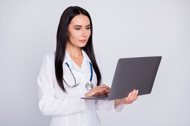 Ragazza concentrata di medico che passa in rassegna le informazioni nel computer portatile su fondo grigio