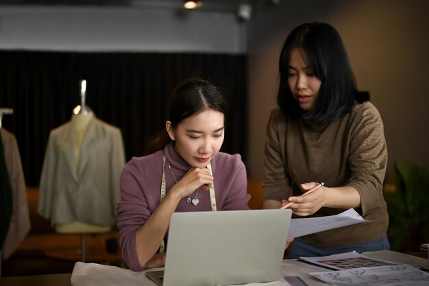 Сосредоточенная портниха, использующая свой ноутбук и работающая со своей помощницей в своей студии.
