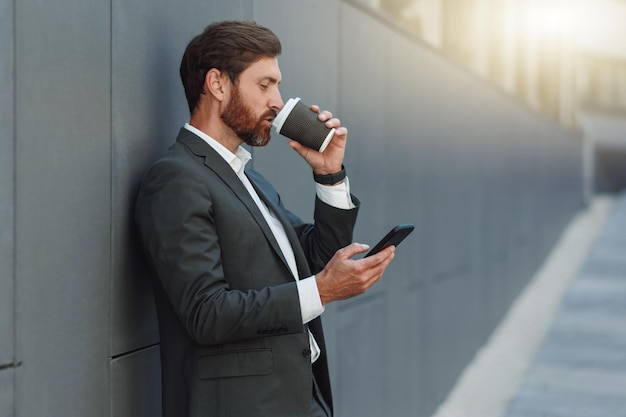 Focused european businessman in suit with phone drinking coffee near office building during break