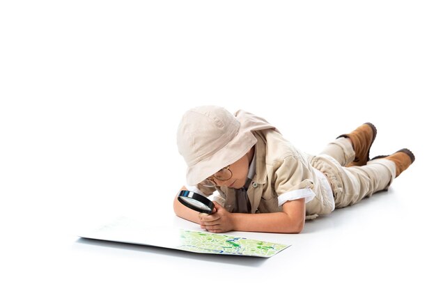 Focused explorer child in hat and glasses looking at map through magnifying glass on white