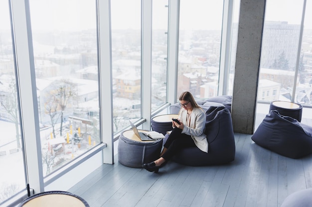 Focused female freelancer sitting in modern workspace with open netbook and checking notifications on mobile phone in casual clothes