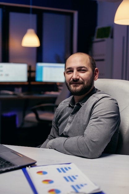 Focused freelancer smiling at camera resting hand on desk late at night. Smart businessman sitting at his workplace in the course of late night hours doing his job.