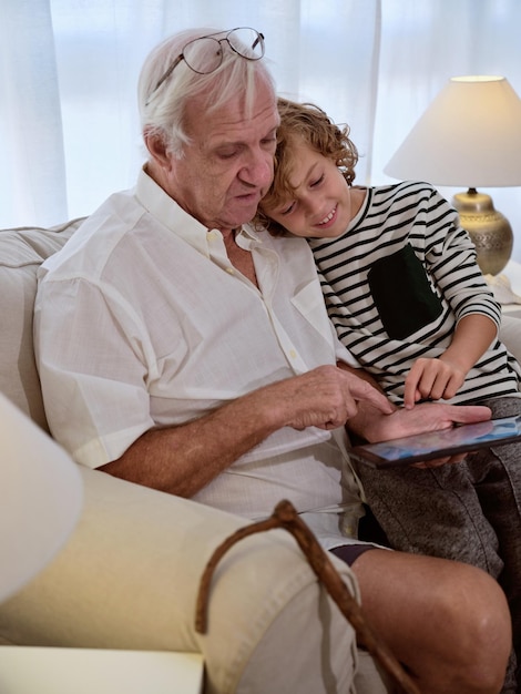 写真 家の明るいリビングルームでソファに一緒に座っている少年と現代のタブレットをサーフィンするカジュアルな服を着た集中した祖父