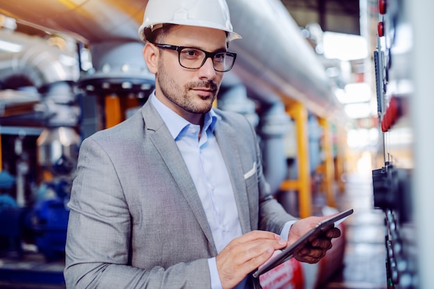 Focused handsome caucasian supervisor in suit and with helmet on head using tablet and looking at dashboard while standing in power plant.