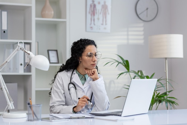 Focused hispanic female doctor sitting at her office desk working on a laptop with paperwork and