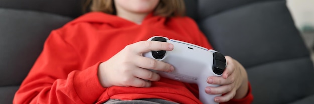 Photo focused junior schoolgirl plays video game at home sitting in armchair little girl in red