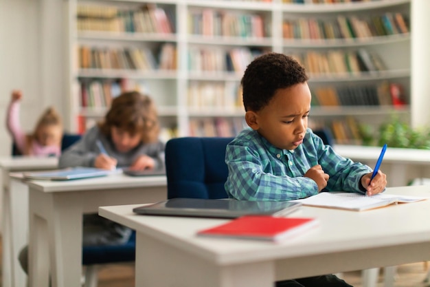 Focused little black schoolboy writing in exercise notebook sitting at desk in classroom with