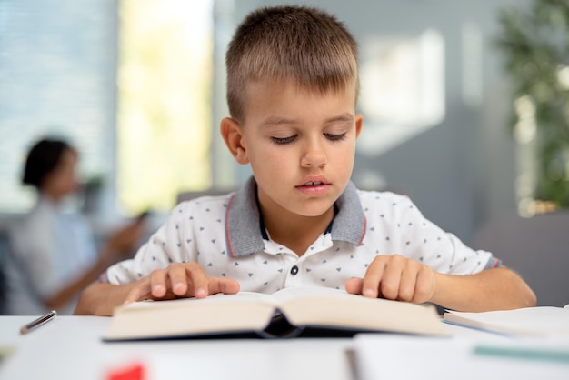 Focused little boy sitting at desk and reading book Caucasian schoolboy spending free time for studying at home Concept of domestic learning