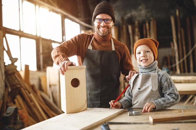 Focused little boy using hammer while making wooden bird house with father in professional joinery workshop