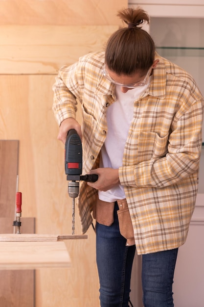 Focused male carpenter drilling hole in wood plank with electric drill while working in home carpent