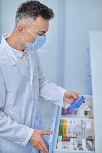 Focused male druggist in a protective mask searching for a medicine in the drawer cabinet