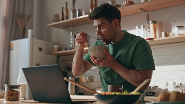 Foto uomo concentrato che fa colazione, legge il portatile, cucina in primo piano, ragazzo che mangia granola.