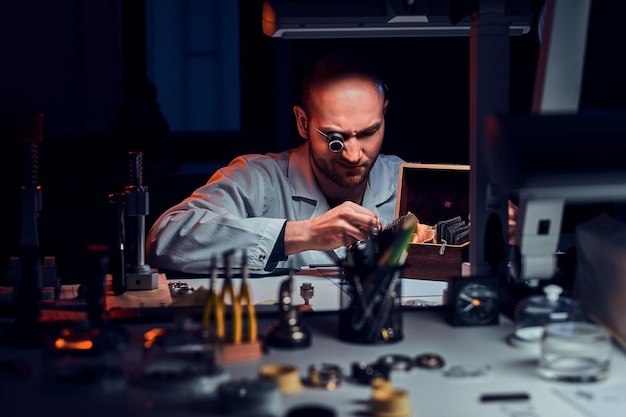 L'uomo concentrato sta lavorando nella sua officina con il monocolo e altri strumenti per riparare vecchi orologi.