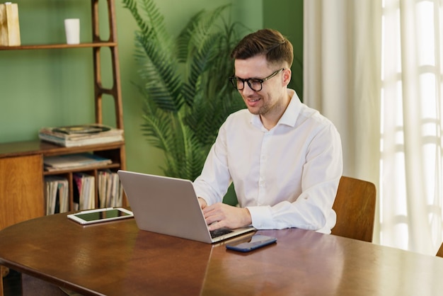 A focused man is working on a laptop with a smartphone and a tablet on a large wooden desk