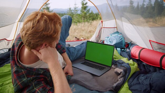 Photo focused man watching movie on laptop with chroma key in tent ginger guy looking video on computer with green screen male hiker relaxing in tourist tent with mountains on background