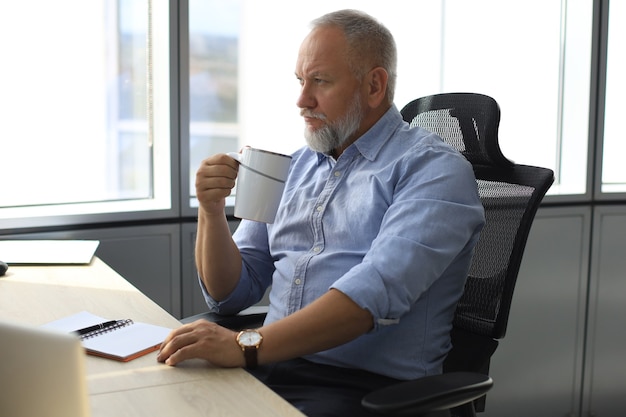 현대 사무실에서 손에 커피 한 잔을 들고 책상에 앉아 있는 동안 깊이 생각하는 성숙한 사업가에 집중했습니다.