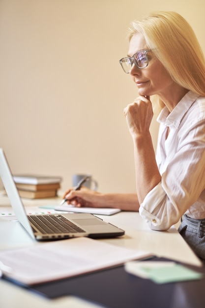 Focused mature businesswoman in glasses looking busy while using laptop making notes working in the