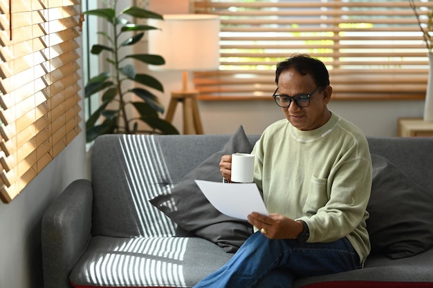 Focused mature man wearing eyeglasses drinking coffee and reading insurance agreement or financial document on couch