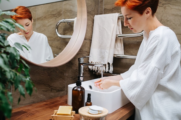 Focused mature woman washing her hands in a stream of water over a sink. She's standing in front of the mirror in a bathroom.