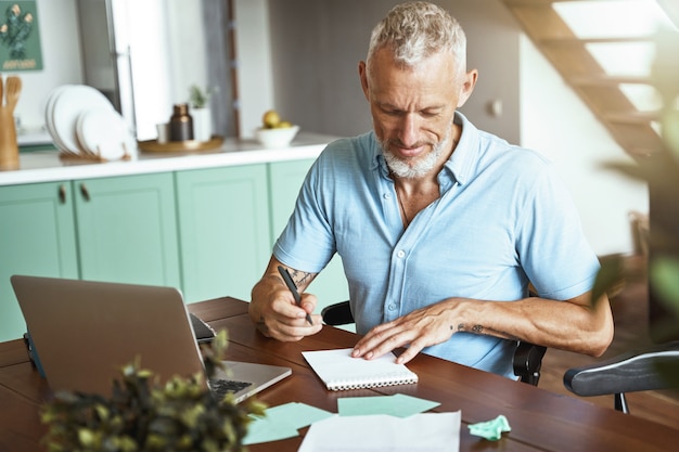 Photo focused middle aged caucasian man making some notes while sitting at table at home during remote