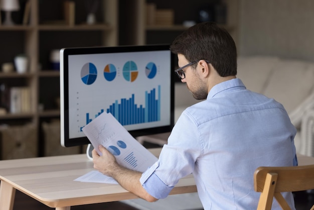 focused millennial business man analyzing marketing reports on desktop monitor reviewing paper graphs financial stats startup project infographics marketer working from home office back view
