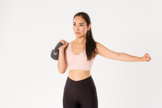 Focused and motivated asian female workout with kettlebell, lift weight and extend one hand, white background.