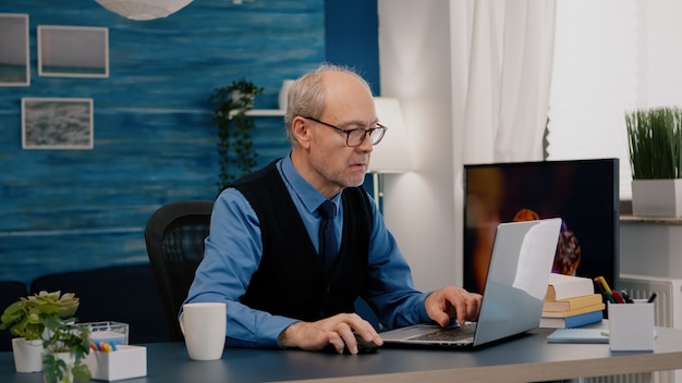 Foto vecchio imprenditore concentrato che controlla la scrittura grafica sul laptop che lavora da casa bevendo caffè in pensione...