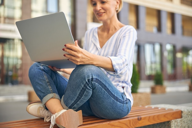 Focused pleased modern female freelancer sitting cross-legged with a laptop on a wooden bench outside