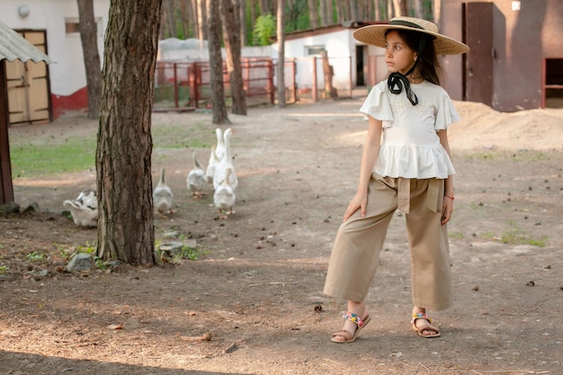 Focused preteen brunette in light blouse stylish culottes and large brim straw hat standing in courtyard of country house located in pine forest looking away full length portrait