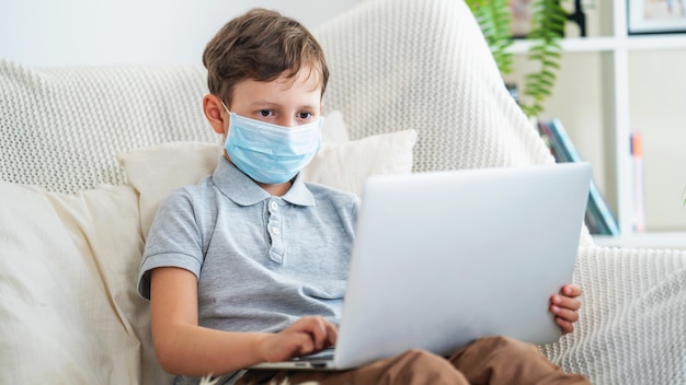 Focused schoolboy in a protective mask is sitting on the sofa with a laptop.