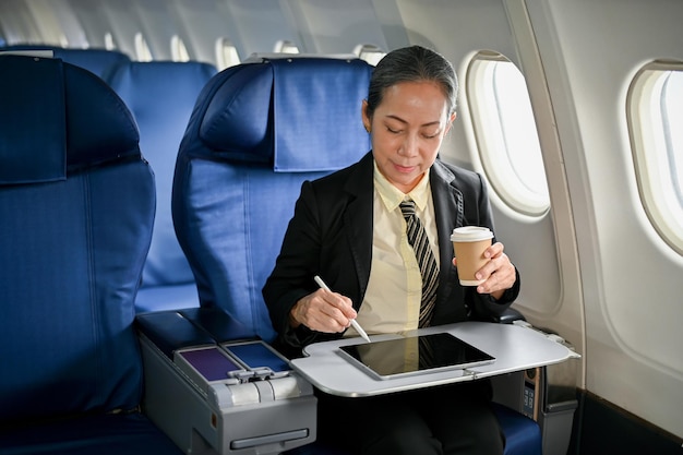 기내에서 커피를 홀짝이며 디지털 태블릿을 사용하는 집중된 아시아계 여성 사업가