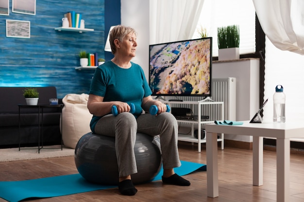 Focused senior woman doing arm exercising using workout dumbbells sitting on swiss ball in living room. Pensioner in sportswear looking at online aerobics training on tablet exercising body resistance