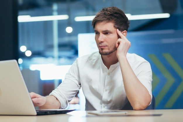 Focused and serious young man working on laptop and with documents in the office at the desk Holds his head He thinks