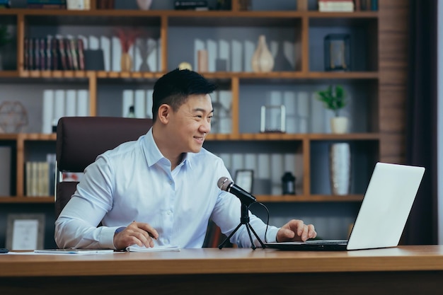 Focused smiling young businessman using professional microphone\
recording audio podcast voice acting or streaming training webinar\
asian looking at laptop screen