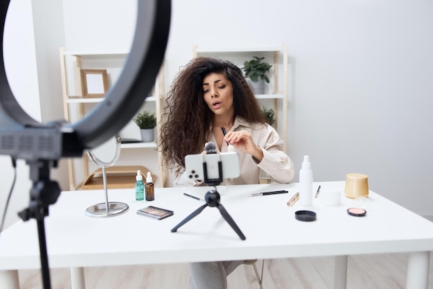 Focused tanned handsome curly latin beauty blogger in linen\
shirt teaches how to use brushes in home white interior copy space\
mockup banner influencer record video review blog using\
smartphone