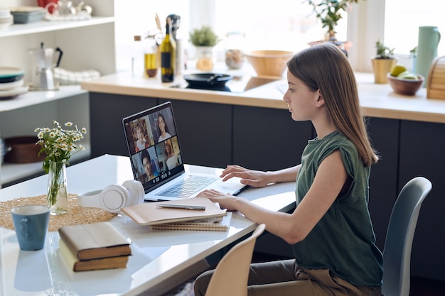 Focused teenage girl sitting at table and using laptop while improving skills at online tutoring class