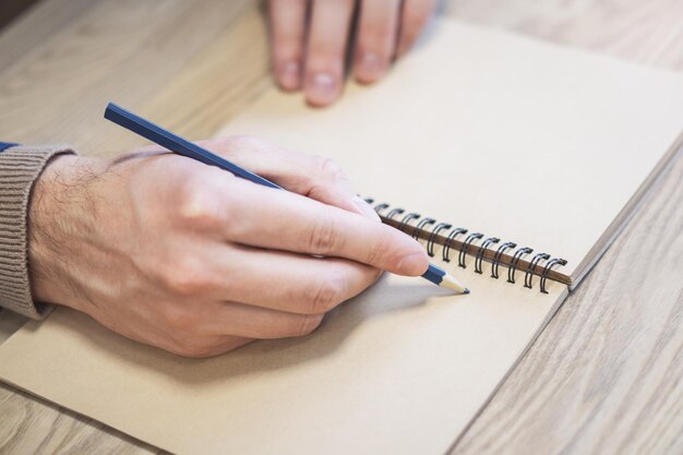 Foto vista focalizzata della mano di un uomo mentre scrive su uno sfondo del blocco note leggermente sfocato