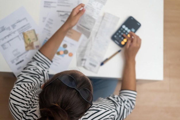 Focused woman counts utility bank bills with calculator of smartphone sitting at desk with documents