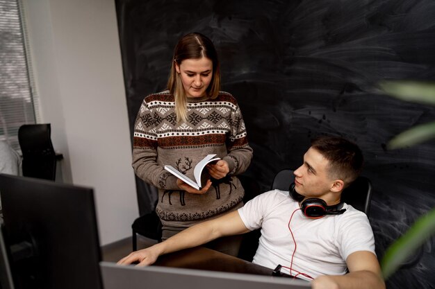 Focused woman holding book in hands and telling something to her man colleague on workplace.