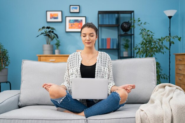 Focused woman smiling relaxed resting on sofa during daily meditation laptop on lap