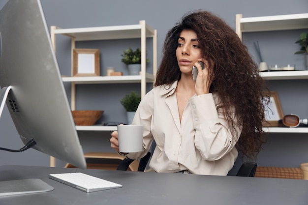 Focused on work tanned adorable curly latin businesswoman in\
linen shirt talk using phone in home office copy space mockup\
banner attractive freelancer work from home using modern desktop\
computer